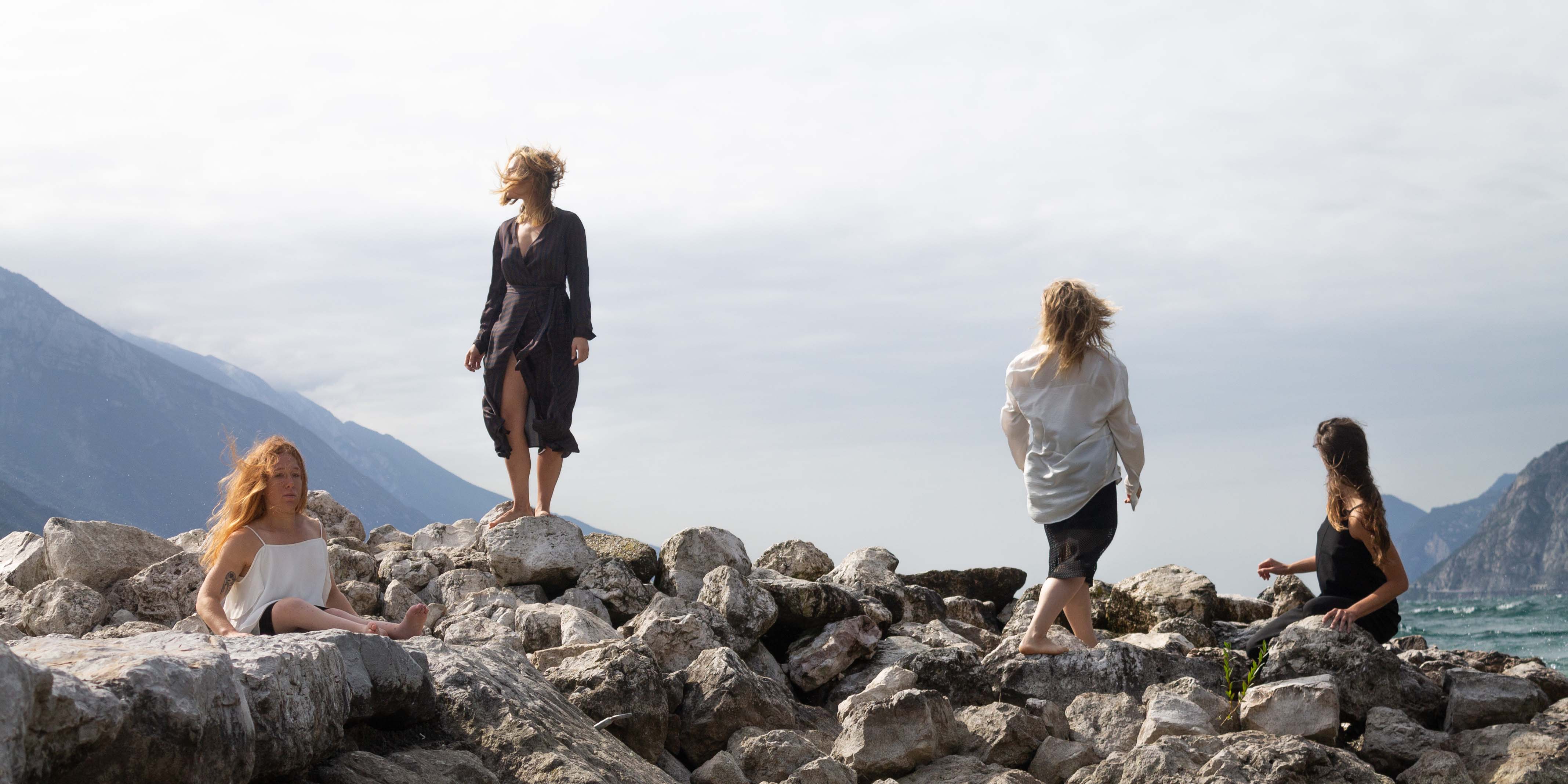 Image for the performance Moby Dick. The image depicts four persons standing and sitting on a pile of rocks, with water and the mountains in the background. The persons are all looking in different directions while the wind pull at their clothes and hair. Photo: Federico Gazza