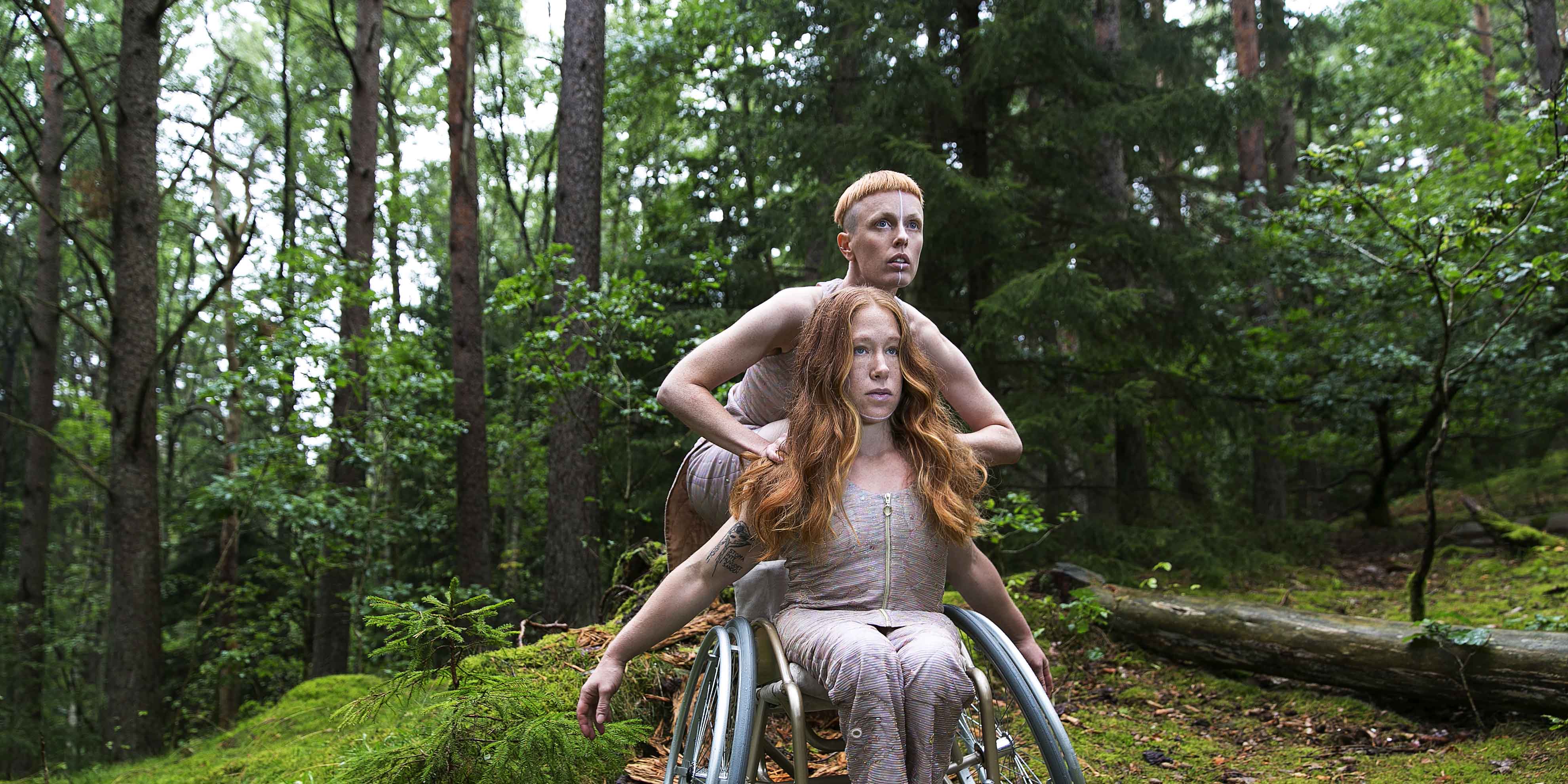 Image for the performance Hannah Felicia. The image depicts the dancers in a forest, wearing costumes shimmering in pink. Felicia is sitting in a wheelchair with her arms pointing backwards, flowing red hair, looking upwards right. Behind her, up on the wheelchair, Hannah crouches, also looking upwards right. Photo: Anna Ósk Erlingsdóttir