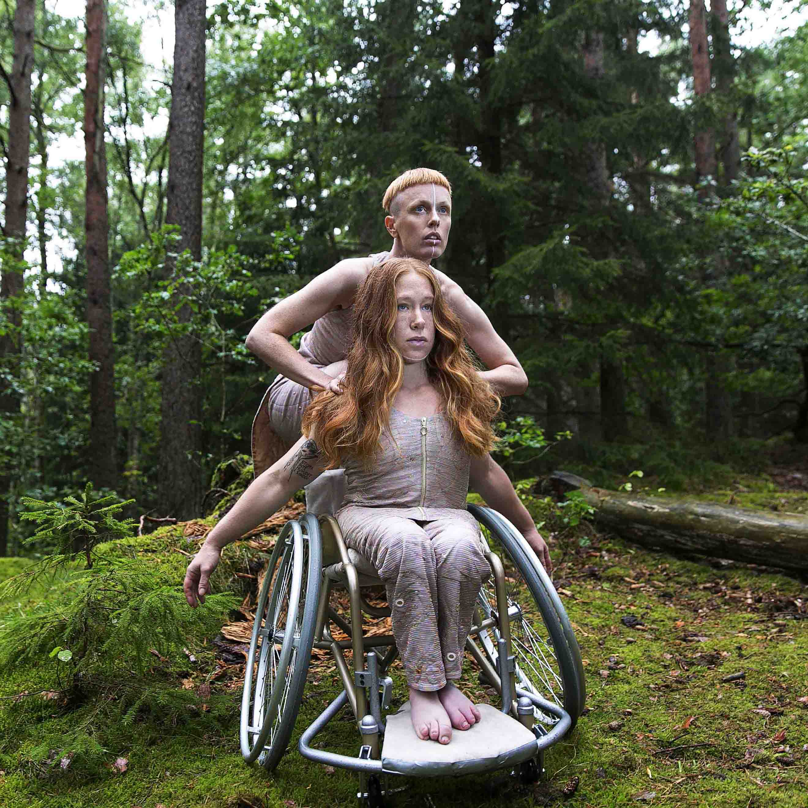 Image for the performance Hannah Felicia. The image depicts the dancers in a forest, wearing costumes shimmering in pink. Felicia is sitting in a wheelchair with her arms pointing backwards, flowing red hair, looking upwards right. Behind her, up on the wheelchair, Hannah crouches, also looking upwards right. Photo: Anna Ósk Erlingsdóttir