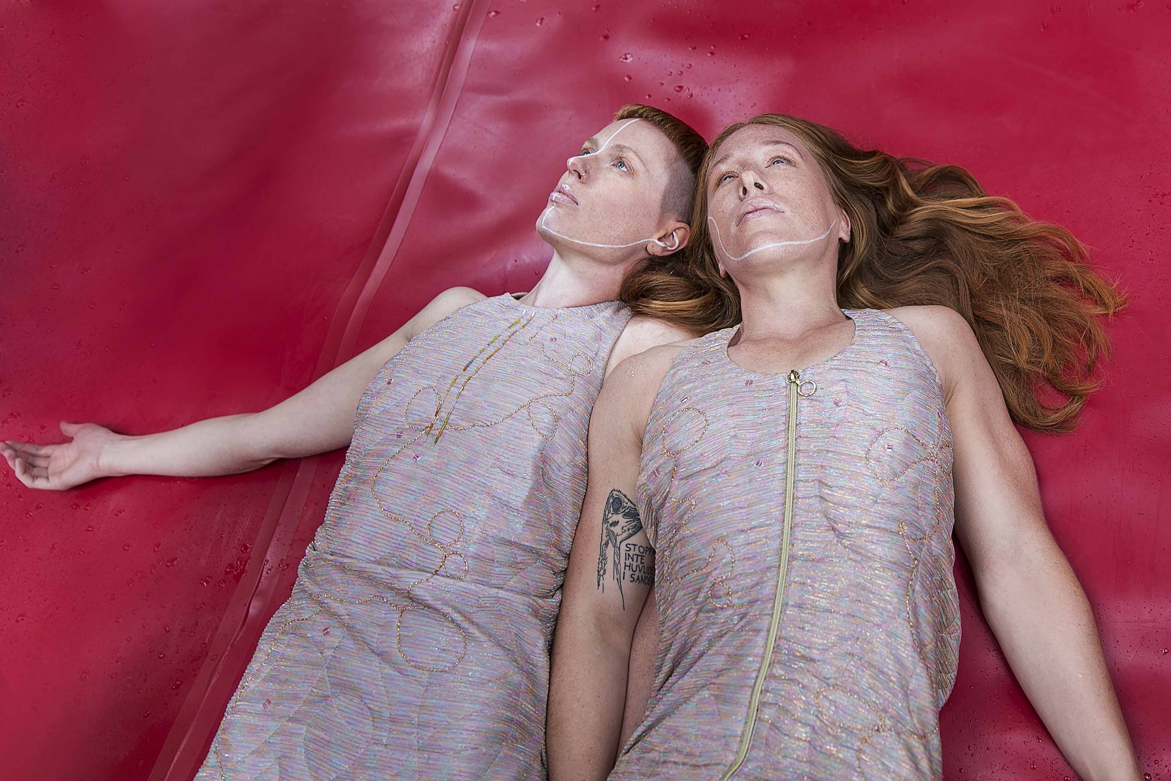The image depicts dancers Hannah Karlsson and Felicia Sparrström who are lying down on a red backdrop, each have one arm outstretched. Both have red hair and are wearing costumes that glitter in pink and silver. Photo: Anna Ósk Erlingsdóttir