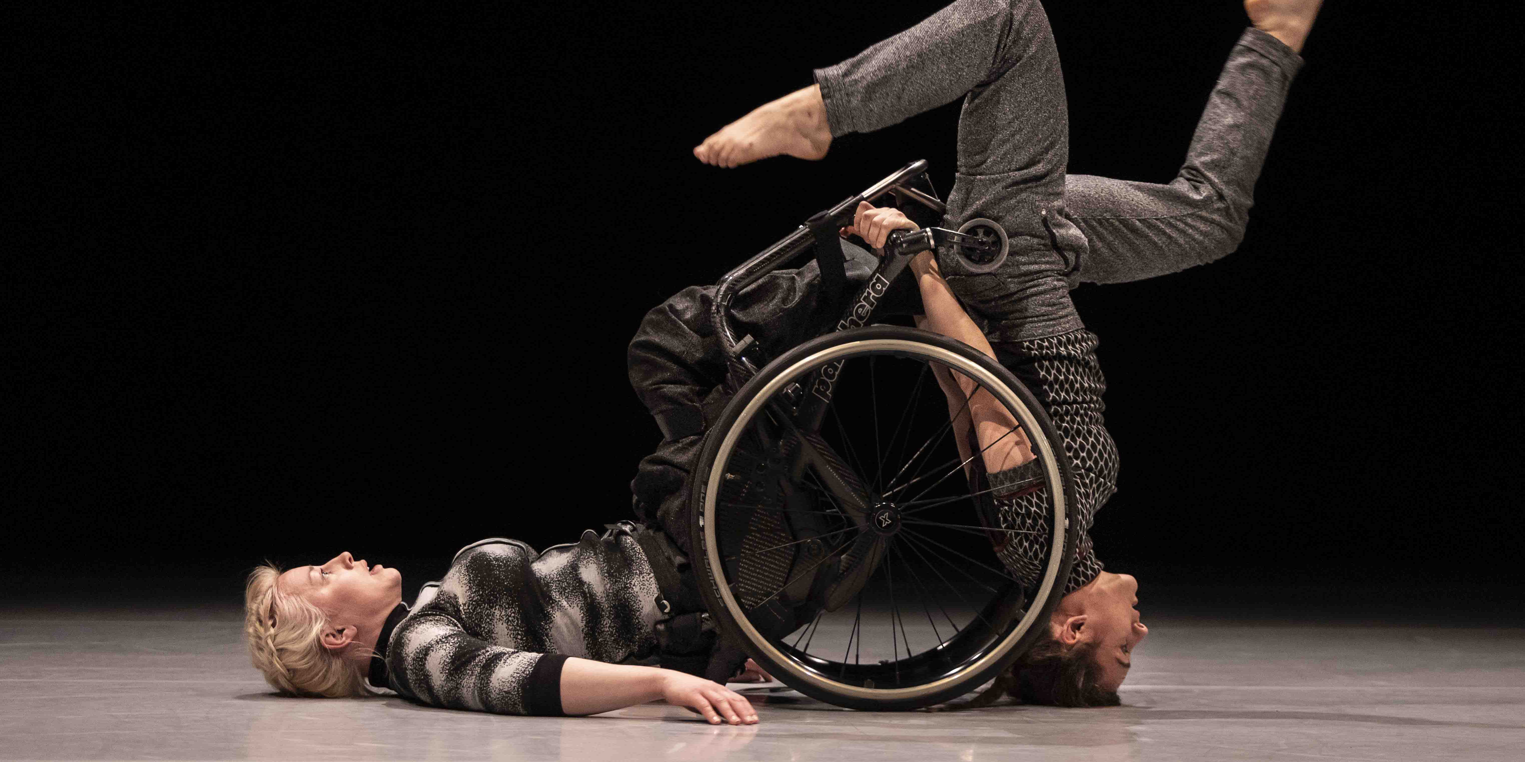 The image depicts two dancers in profile against a black background. The dancer on the left lies on her back with her legs up on a wheelchair that has tipped over. The dancer on the right stands on her head with spread legs, while holding onto the wheelchair with her hands. Photo: Tilo Stengel