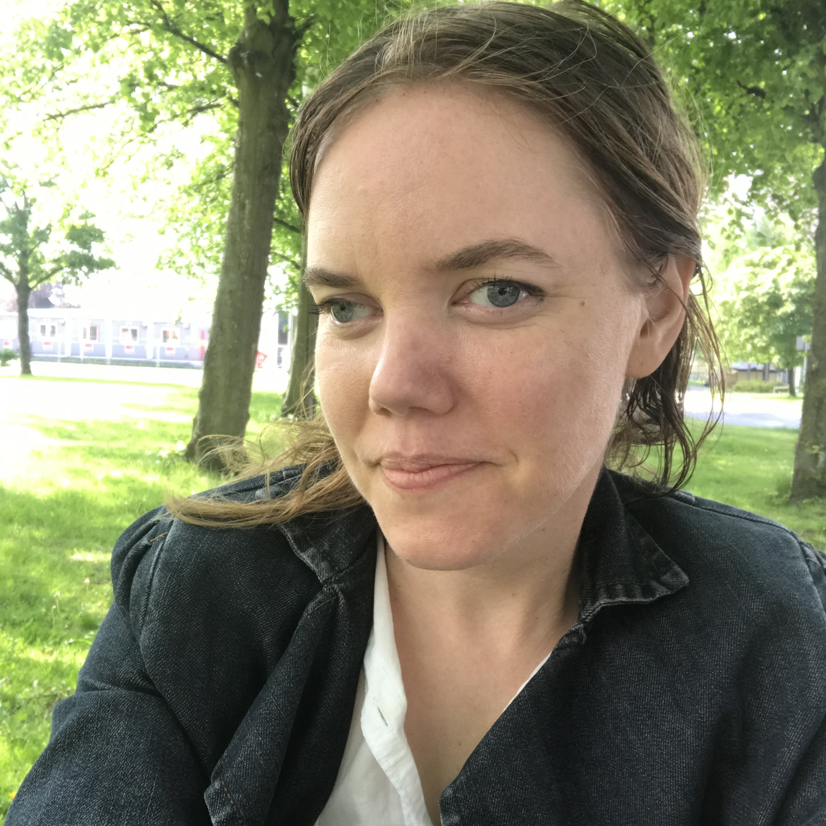 The picture shows dancer Ylva Andersson. Ylva is seen at the front of the picture. She is wearing a black denim jacket and a white shirt. In the background we see a green park where the sun finds its way through the trees.