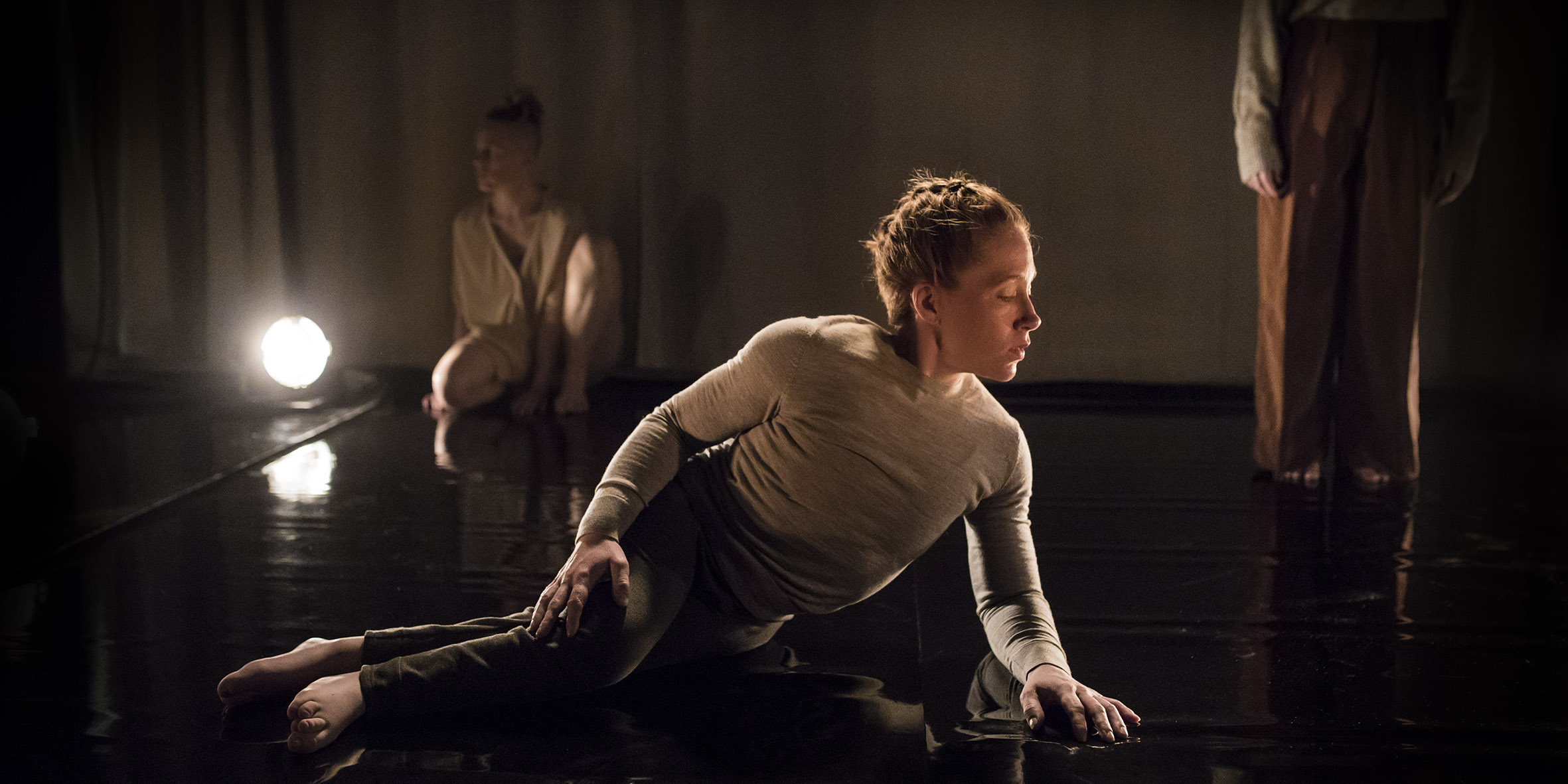 In the foreground lies a dancer with her legs to the left and her upper body to the right. She has braided red hair and are looking down at the floor. In the background to the left sits a dance out of focus next to a bright spotlight. In the background to the right we see the legs and parts of the arms of a person. The picture is dark and light is reflected on the shiny floor. Photo: Polina Ulianova