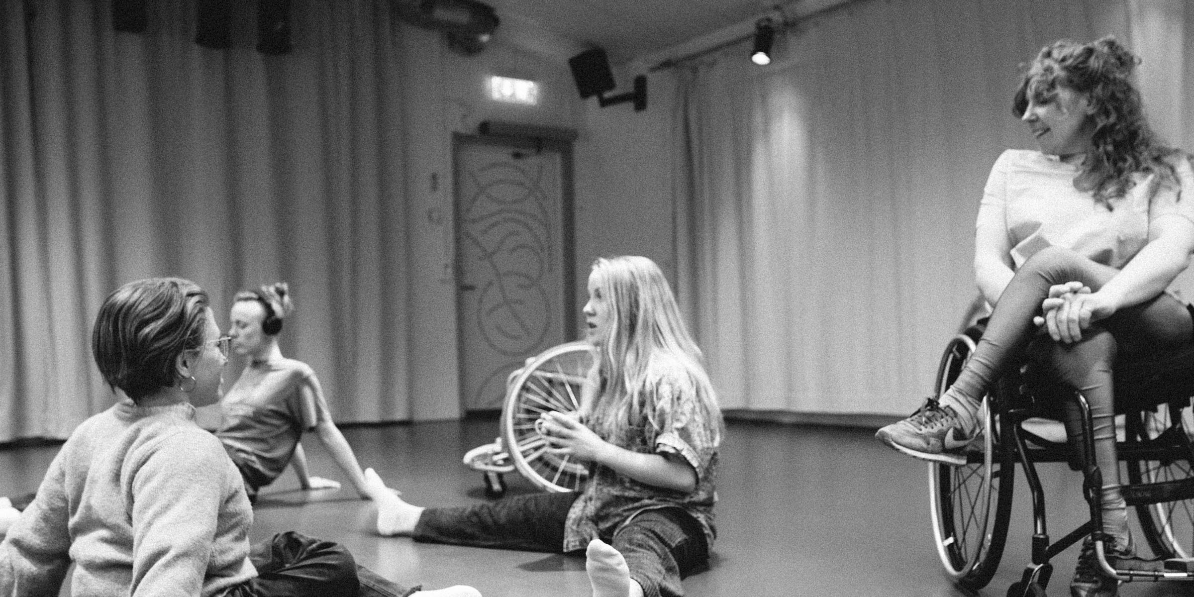 A black and white image depicting four dancers. Three of them are sitting on the floor and the forth one in a wheelchair. In the background there are curtains and an additional wheelchair. Photo: Malin Johansson