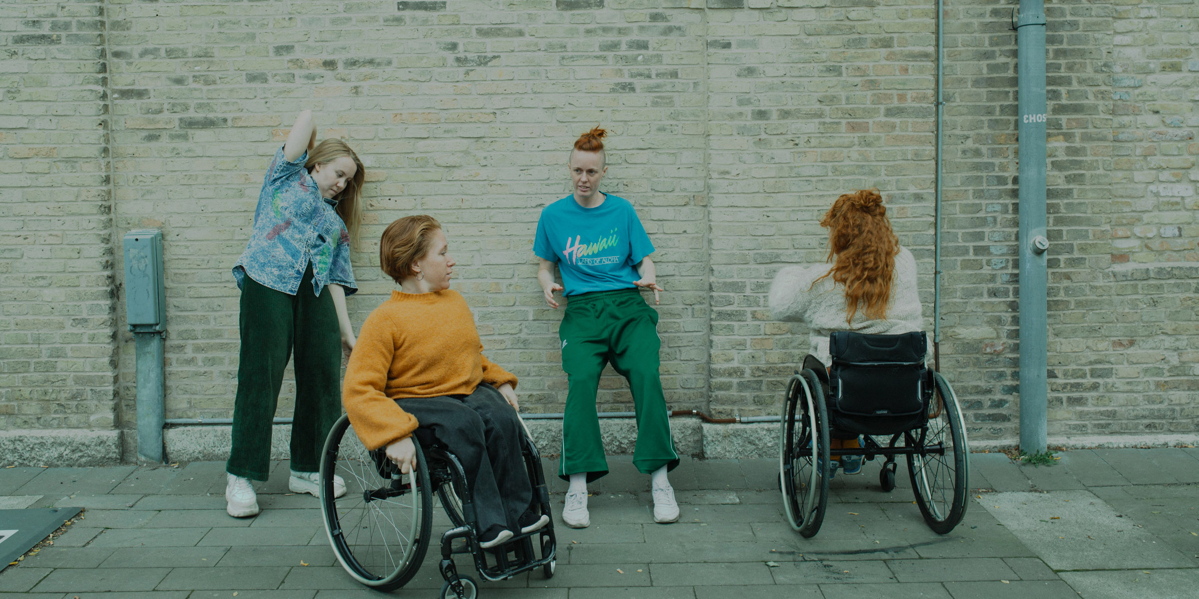 Four dancers, outdoors by a brick wall. Two dancers stands leaning against the wall. One sits in a wheelchair with her back to the camera. One sits in a wheelchair with some distance from the wall, looking at one of the other dancers. Photo Malin Johansson