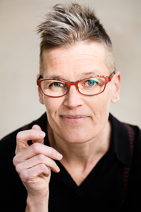 Porträttbild av Veera Suvalo Grimberg, konstnärlig ledare. Veera har gråbrunt kort hår, röda glasögon, svart skjorta med rödbruna hängslen och sin högra hand halvsluten. Foto: Polina Ulianova