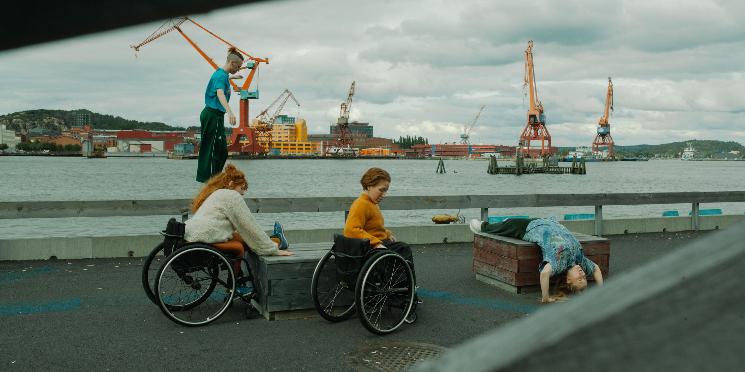 In the picture we see four dancers. They are outdoors. On a wharf by a river. There are cranes in the background. Two dancers sit in wheelchairs, one of whom has one leg up on a wooden box. A third dancer lies on another wooden box and a fourth dancer balances on a concrete edge. Photo Malin Johansson