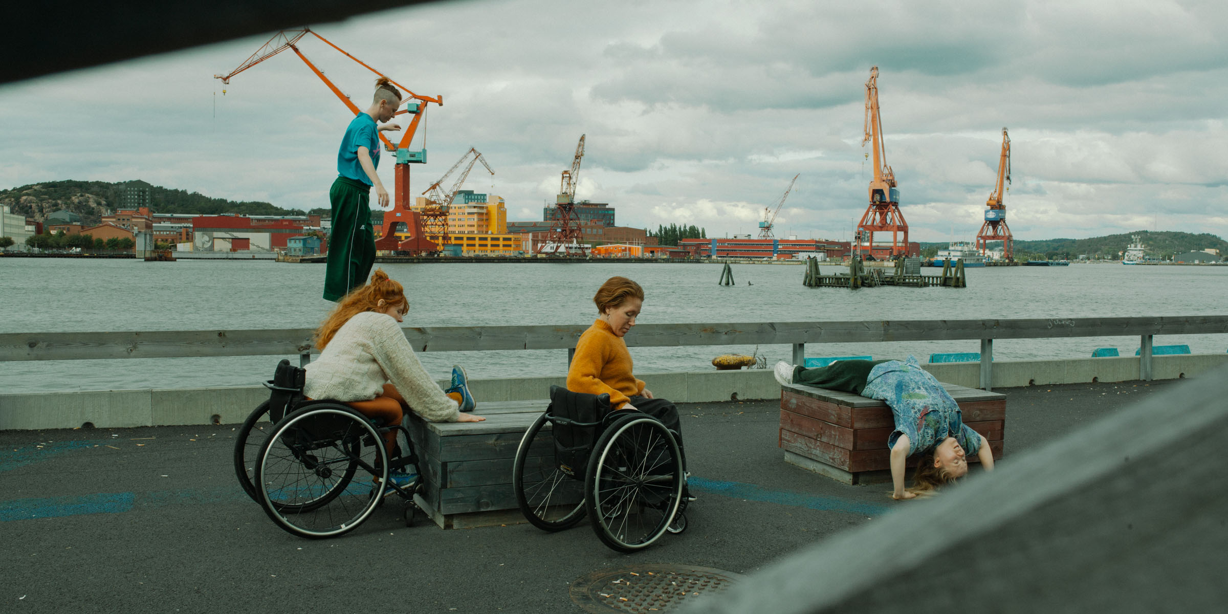 På bilden ser vi fyra dansare. De är utomhus. På en kaj vid en älv. Det är lyftkranar i bakgrunden. Två dansare sitter i rullstol, varav en har ena benet upp på en trälåda. En tredje dansare ligger på en annan trälåda och en fjärde dansare går balansgång på en betongkant. Foto Malin Johansson