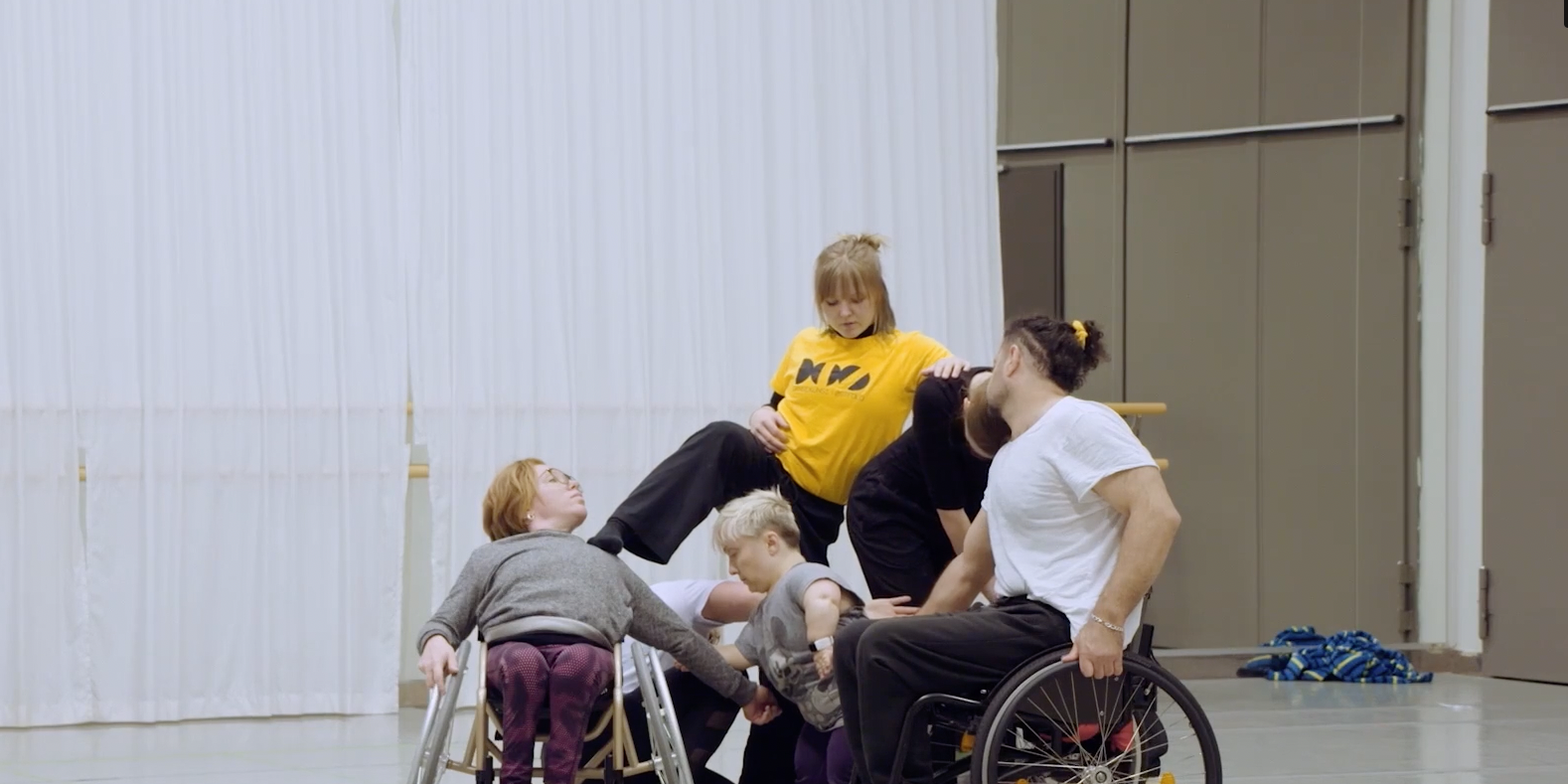 The image depicts five dancers in a dance studio standing close together. Three of them are standing up, two are sitting in wheelchairs. They all look in different directions.
