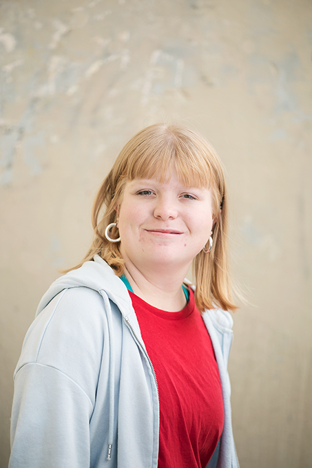 Porträttbild av dansaren Ida Häggström. Ida har blont hår med uppklippt lugg, ringar i båda öronen och en röd t-shirt med en vit munkjacka över. Foto: Polina Ulianova