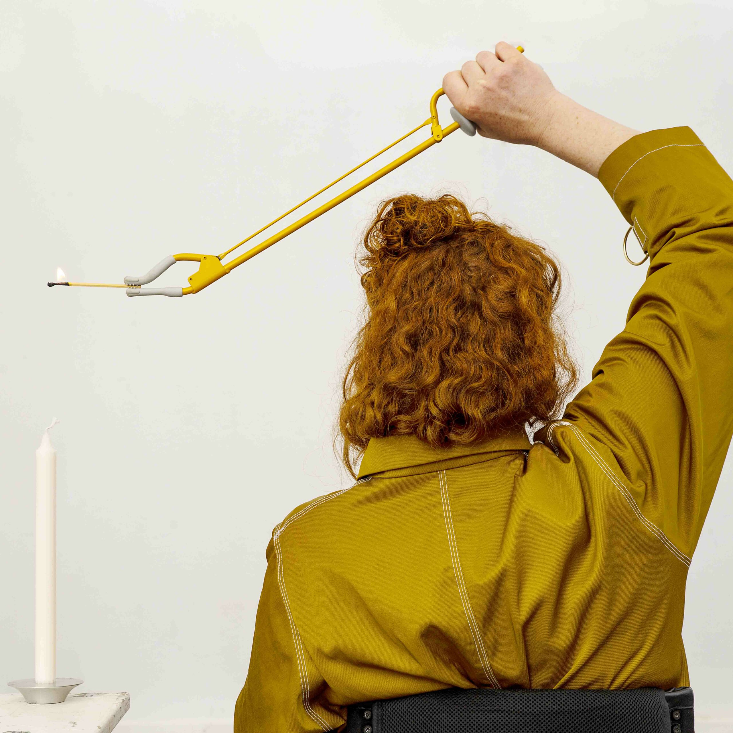 The motion depicts a person with red hair sitting with their back to us, wearing a mustard yellow shirt. In their hand holds a yellow gripper. In the gripper is a lit match above a candle. Photo: Lars Dyrendom