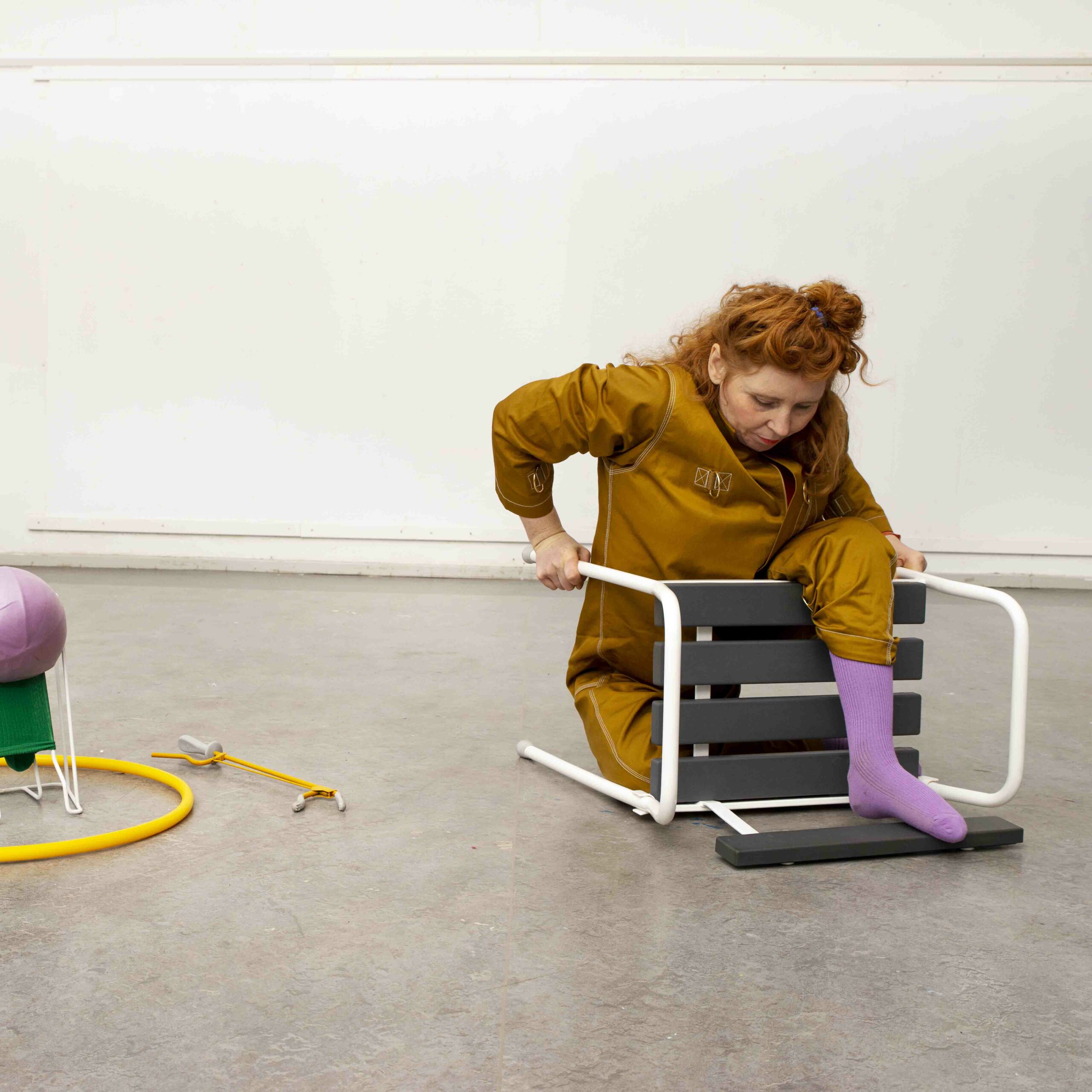 The image depicts a woman wearing a mustard-yellow dress and red hair, trying to climb over a char that been tipped over. To the left we see a yellow hula hoop, a yellow gripper, a purple ball and a green sock. Photo: Lars Dyrendom