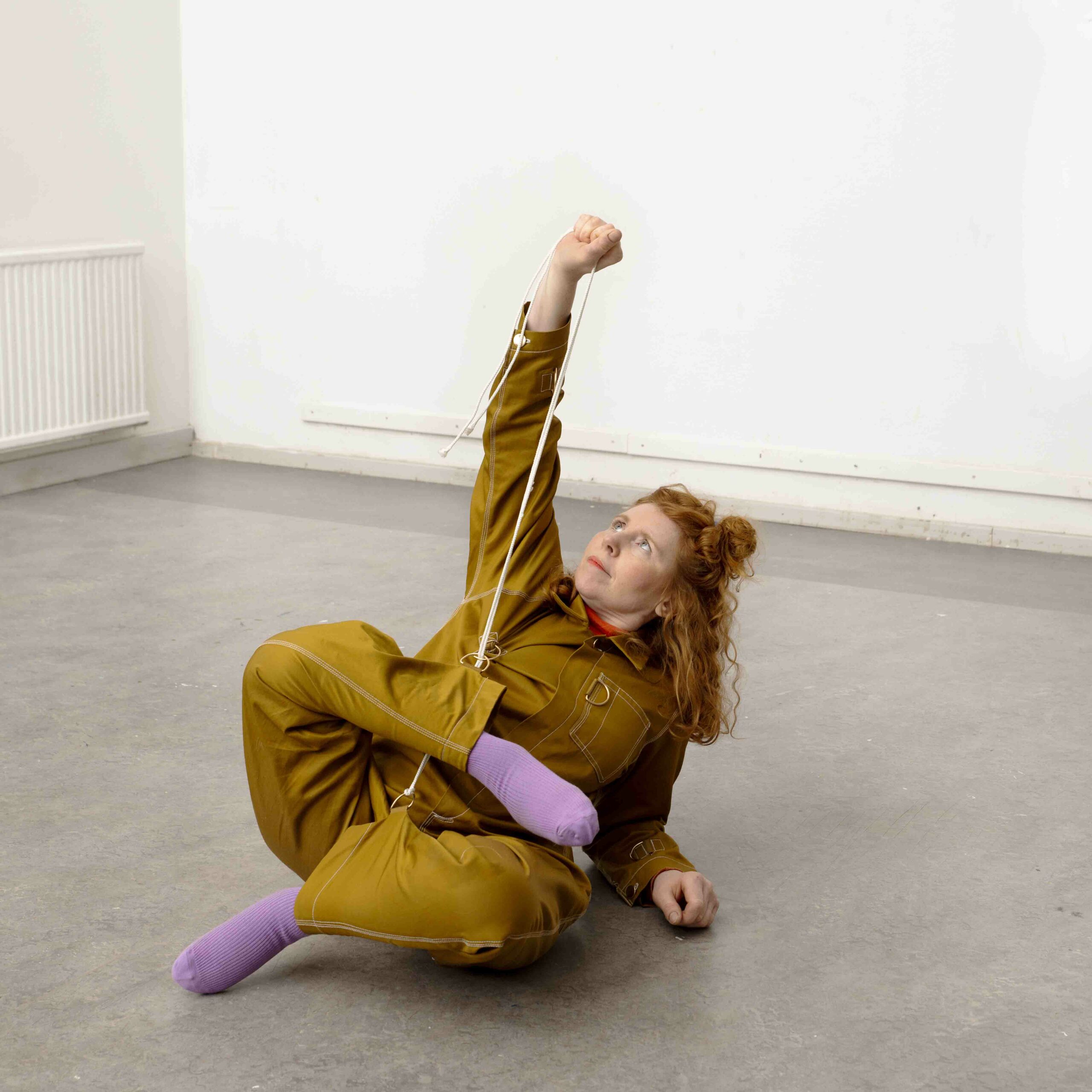 Bilden visar en kvinna iklädd en senapsgul dräkt och rött hår som ligger på golvet samtidigt som hon håller upp ett av sina ben med ett snöre. Foto: Lars Dyrendom