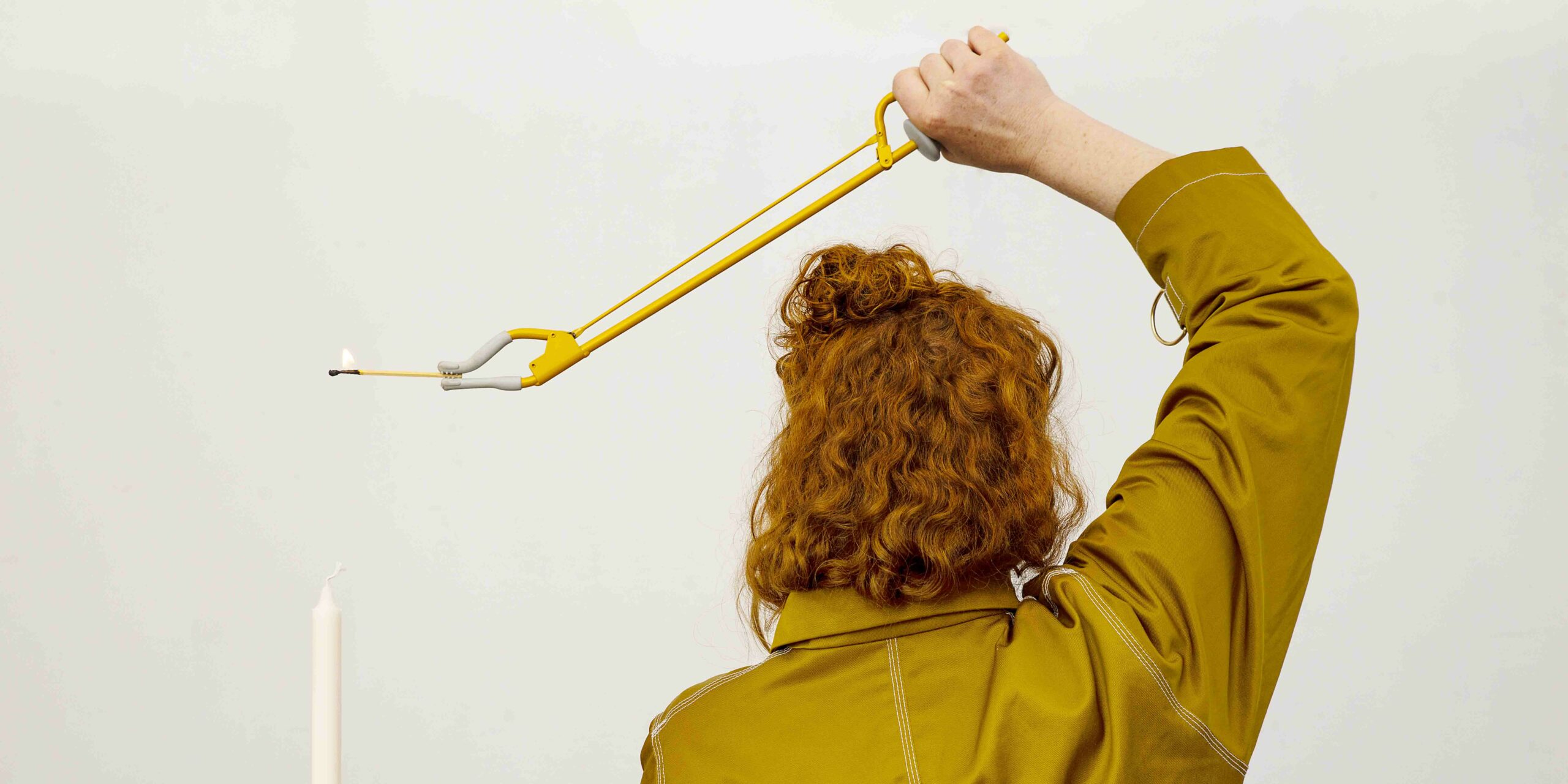 The image depicts a person with red hair sitting with the back towards us, wearing a mustard-yellow shirt. In their hand is a yellow gripper holding a lit match above a stearic candle. Photo: Lars Dyrendom