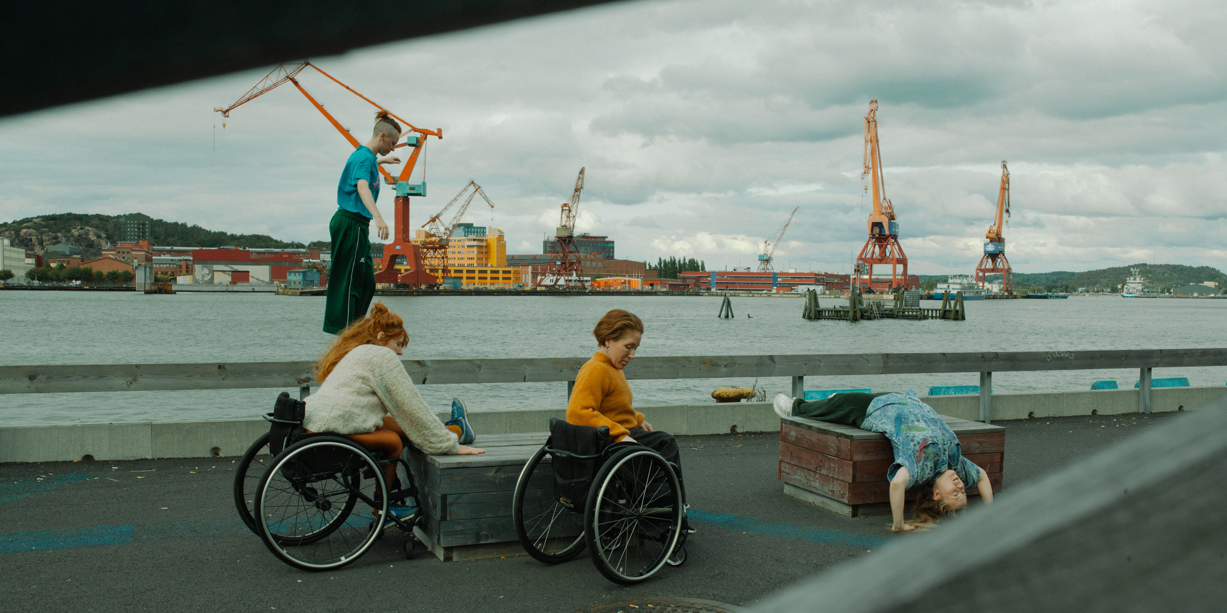 In the picture we see four dancers. They are outdoors. On a wharf by a river. There are cranes in the background. Two dancers sit in wheelchairs, one of whom has one leg up on a wooden box. A third dancer lies on another wooden box and a fourth dancer balances on a concrete edge.Photo Malin Johansson