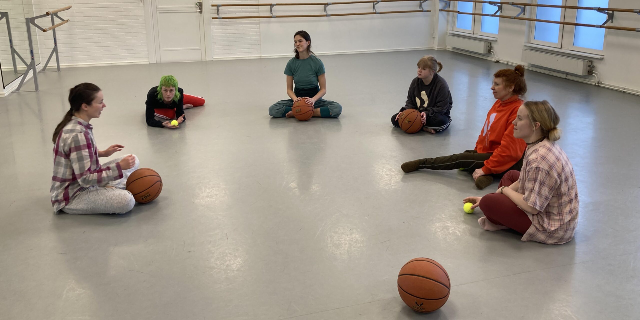 5 dansare sitter på golvet i en dansal. De sitter i en större cirkel och pratar med en workshopledare som också sitter. Workshopledaren har en basketboll i famnen, det har också 2 av dansarna. En basketboll ligger på golvet. Foto Veera Suvalo Grimberg