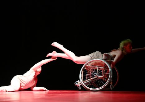 Two female dancers in profile looking to the right. They are on a stage with a black background and a red dance mat. One dancer lies on the floor with his chest upright and extends one arm towards the other dancer who lies on a wheelchair with one arm extended to the right. Photo Sandro DondaL