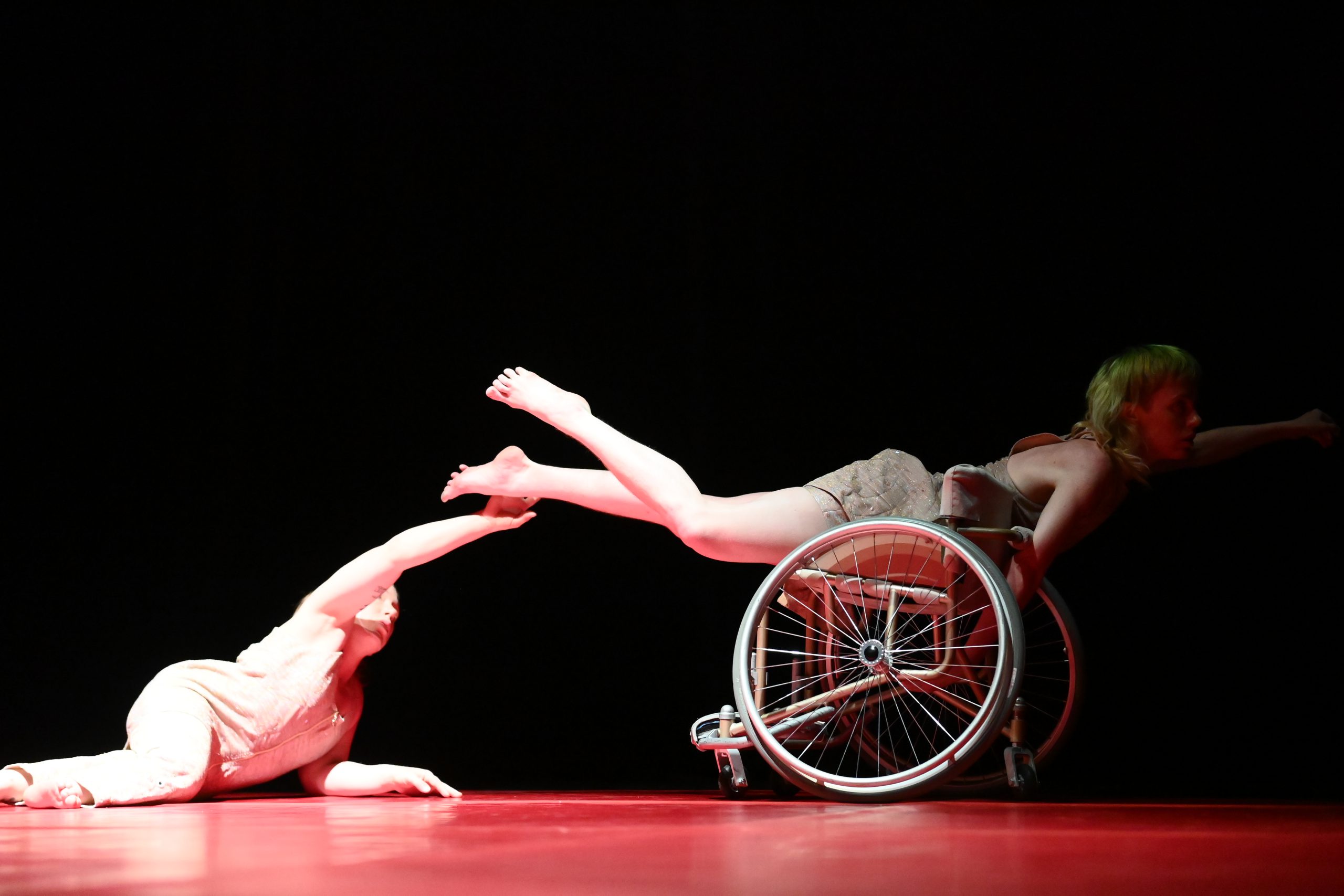 Two female dancers in profile looking to the right. They are on a stage with a black background and a red dance mat. One dancer lies on the floor with his chest upright and extends one arm towards the other dancer who lies on a wheelchair with one arm extended to the right. Photo Sandro DondaL
