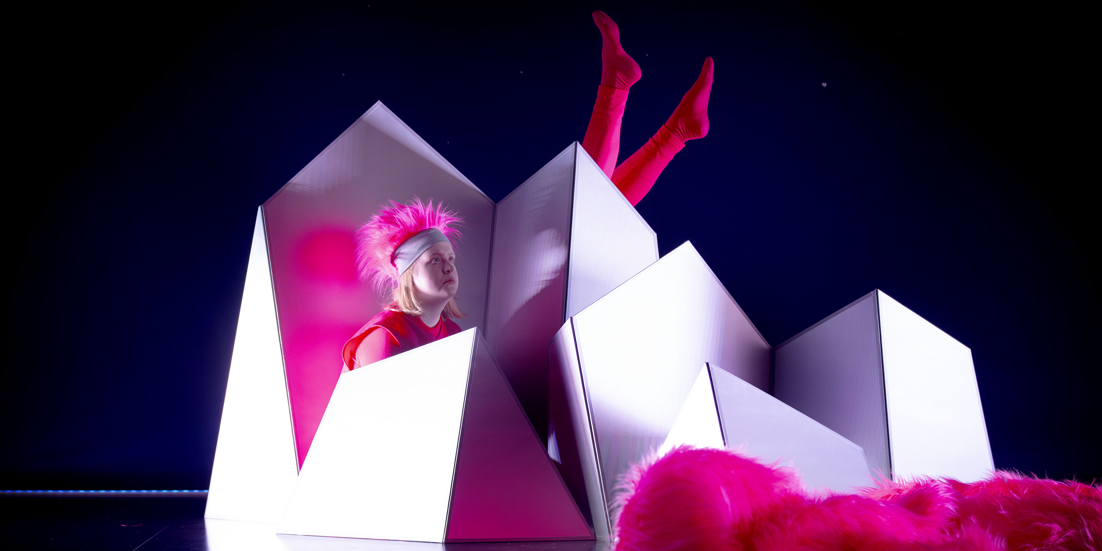 A dancer sits between a group of white screens with pointy edges, resembling icebergs. We see the dancer from their shoulder up, with their gaze fixed on the viewer. The dancer is wearing a pink body stocking with a vest on top. On their head, the dancer wears a hat with fluffy pointy pink hair. To the right behind the dancer, we see two pink legs coming up. The background is dark blue and in the foreground, we see a large fluffy pink tail. Photo: Malin Arnesson