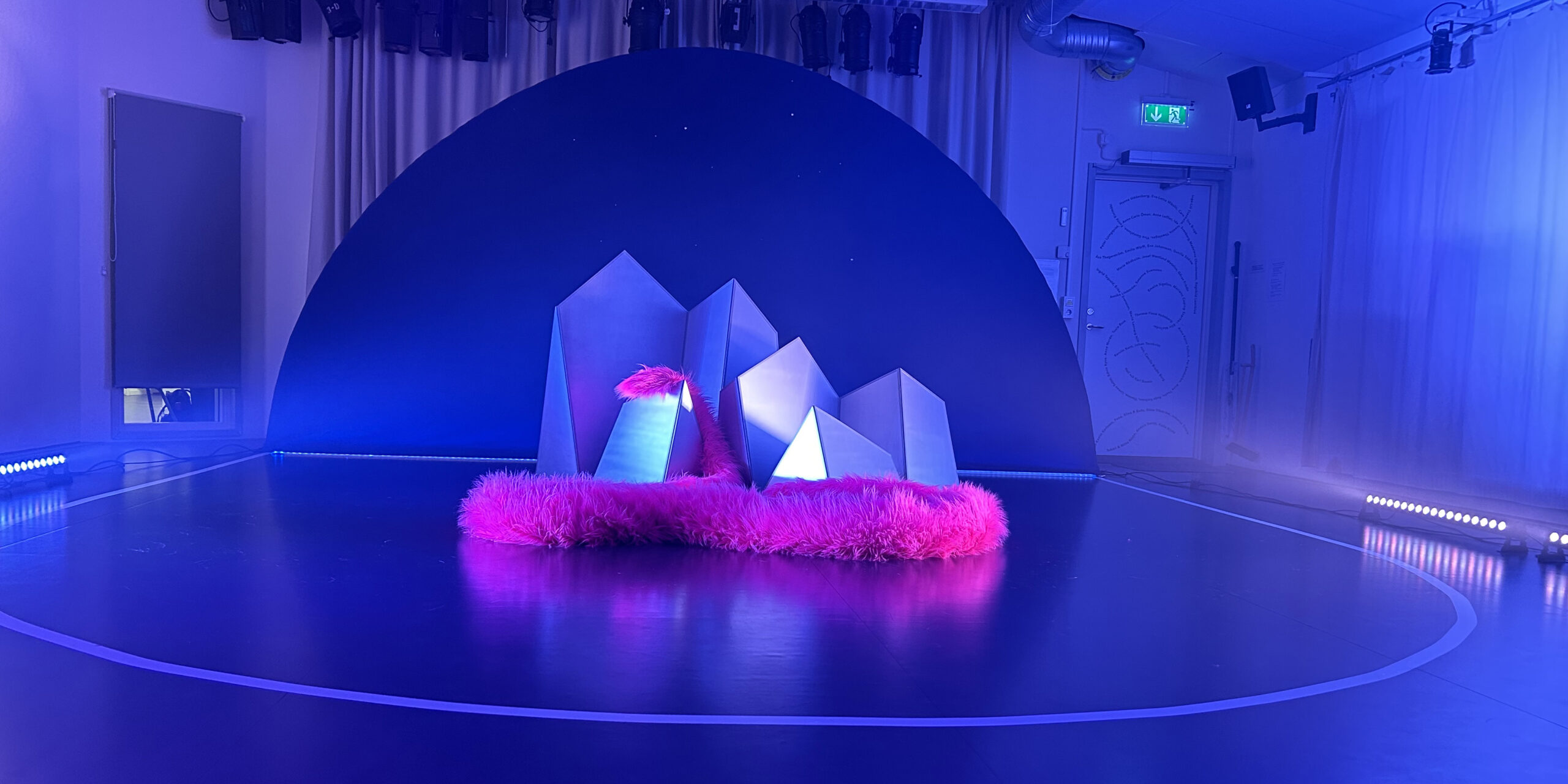 A room in dark blue light. In the center of the image, there are white screens resembling mountains. Between the screens, a large, fluffy cerise-colored tail winds its way. Behind the mountains, there is an oval-shaped dark blue background. Photo by Hedda Gullander. Set design by Karin Agélii.