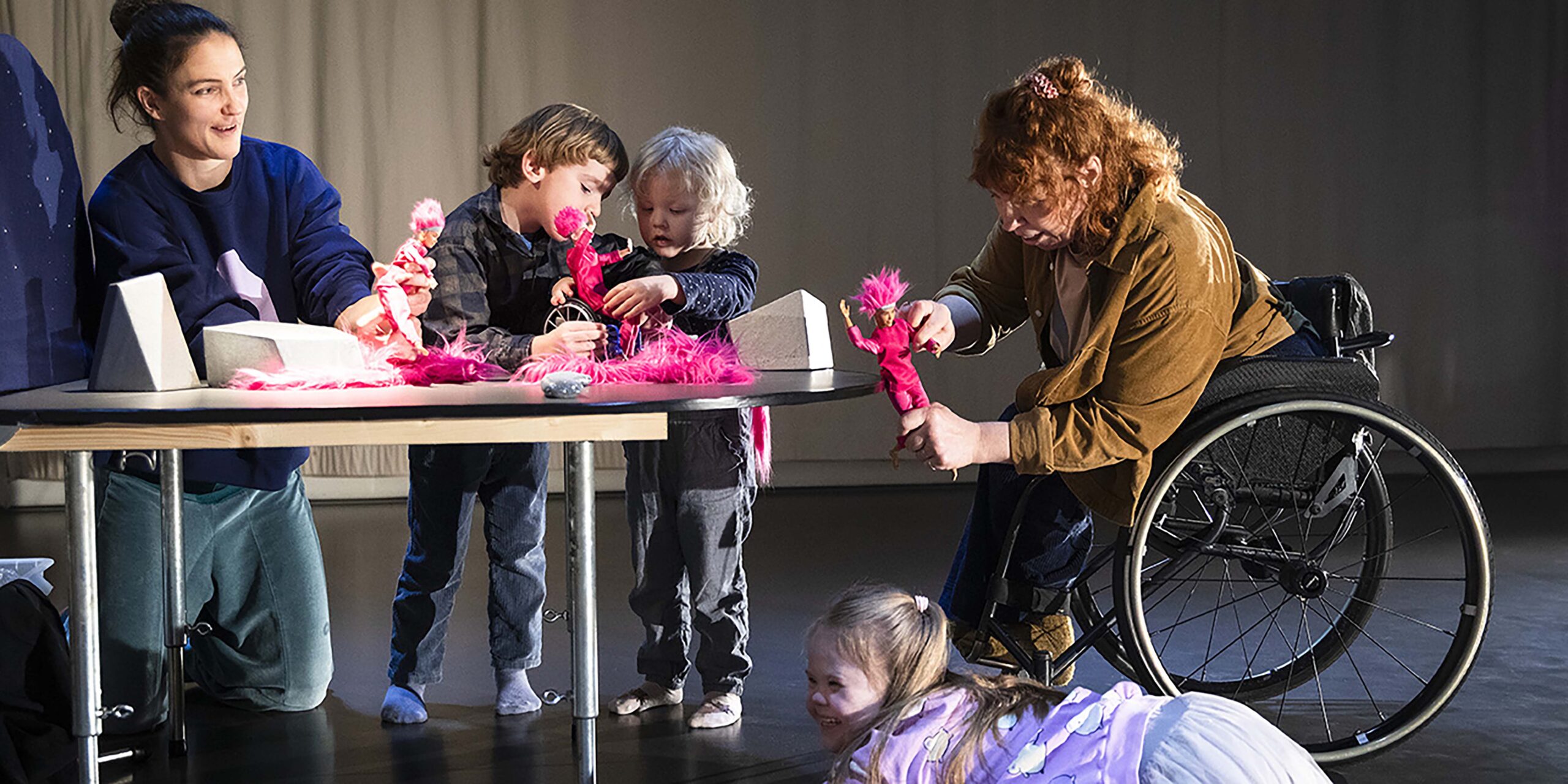 Two dancers sitting at a table. Two children standing by the table. The dancers and the children are exploring and playing with dolls dressed in pink clothes and pink fluffy hats. A third child is lying on the floor next to the table. The child is looking diagonally to the left and laughing. Photo Lina Ikse.